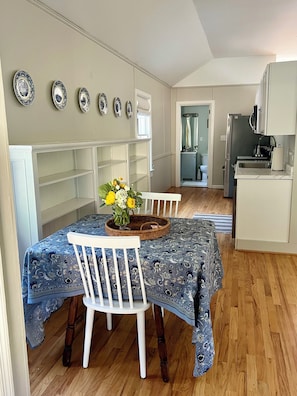Un comedor con una mesa dispuesta para dos personas, una silla blanca y un florero con flores. Se observa una cocina al fondo, con un refrigerador y un microondas.