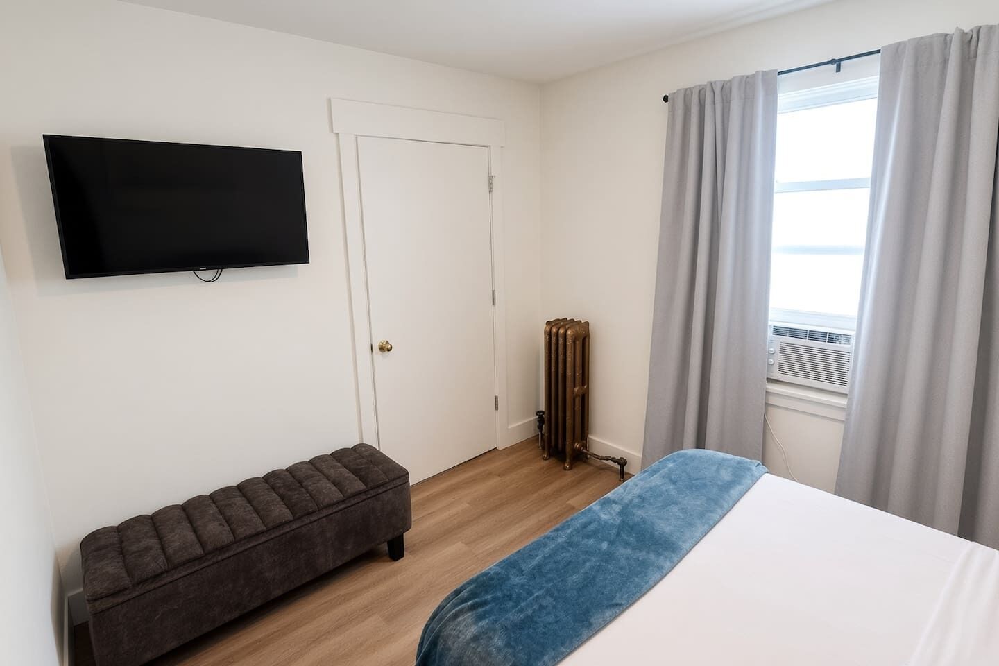 Quarto com cama, um banquinho, radiador, televisão e janela com cortinas.