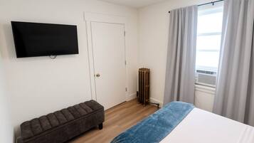 Quarto com cama, um banquinho, radiador, televisão e janela com cortinas.