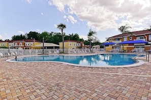 A swimming pool with lounge chairs and umbrellas, surrounded by a brick patio and buildings in the background. - Spacious 3-Bedroom TH Disney (Kissimmee)