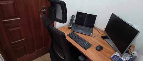 A wooden desk with a laptop, keyboard, mouse, and a monitor. There is a black office chair and a wooden door in the background.