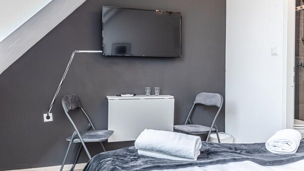 A modern hotel room with a bed, a desk, two chairs, a flat-screen TV mounted on the wall, and a shower area.