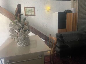 A room with a dining table, a vase of flowers, a wooden chair, a black sofa, a wall-mounted lamp, and a framed picture.
