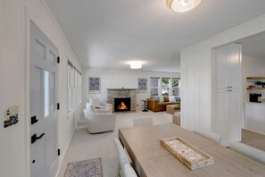 Uma sala de estar ampla com lareira, uma área de jantar com uma mesa comprida e uma cozinha com bancada e armários.