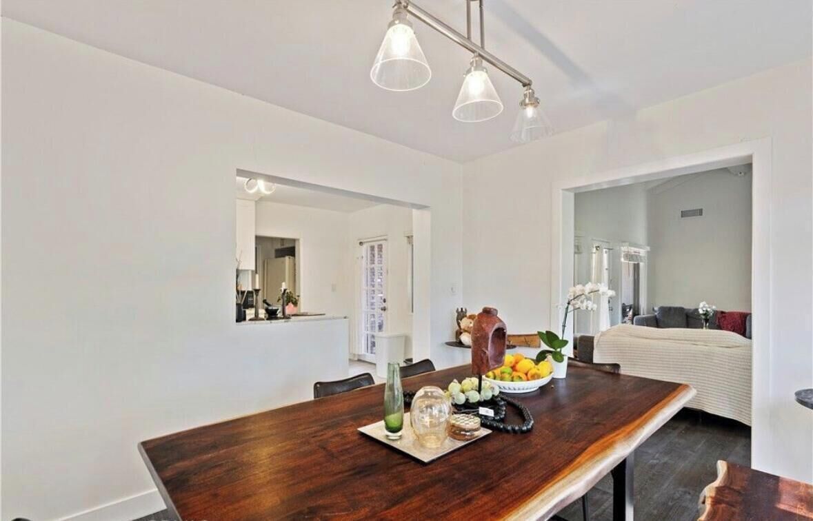 A modern dining area with a wooden table, a bowl of fruit, and a vase. There is a mirror on the wall and a doorway leading to another room.