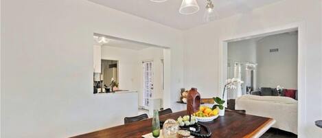 A modern dining area with a wooden table, a bowl of fruit, and a vase. There is a mirror on the wall and a doorway leading to another room.