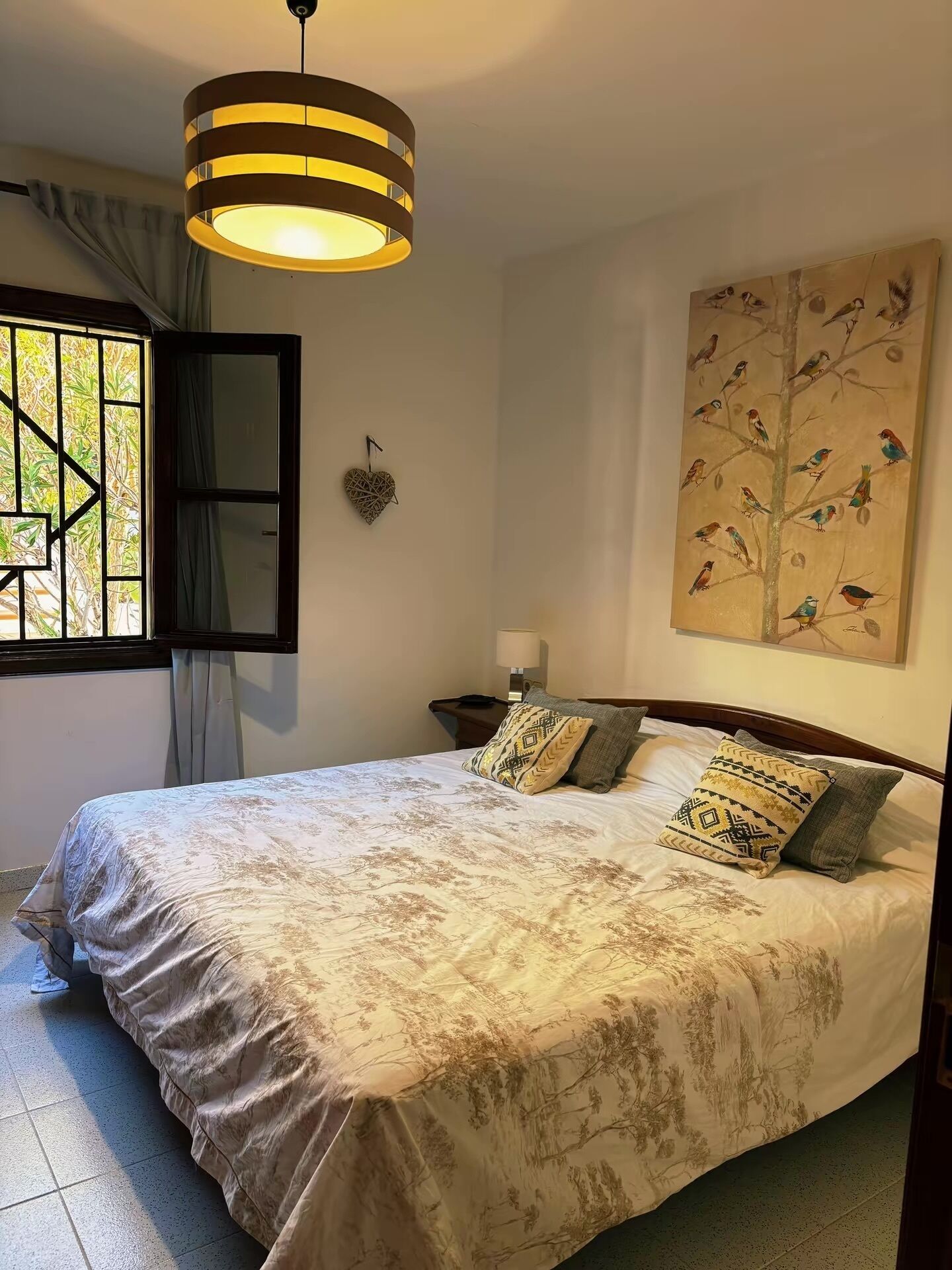 A bedroom with a bed, a hanging light fixture, a window with curtains, a wall-mounted heart decoration, and a bird-themed painting.