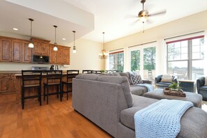 A spacious living area with a grey sofa, wooden flooring, and a kitchen with wooden cabinets and modern appliances.