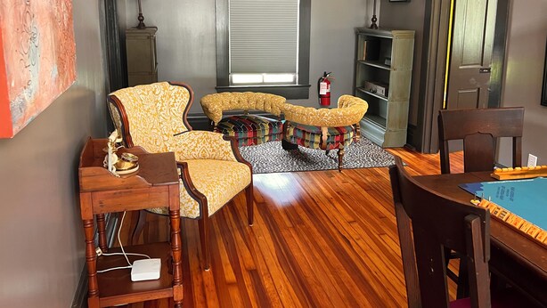 Un soggiorno con pavimento in legno, una poltrona gialla con motivi decorativi, un poggiapiedi multicolore, un tavolino in legno e una libreria.