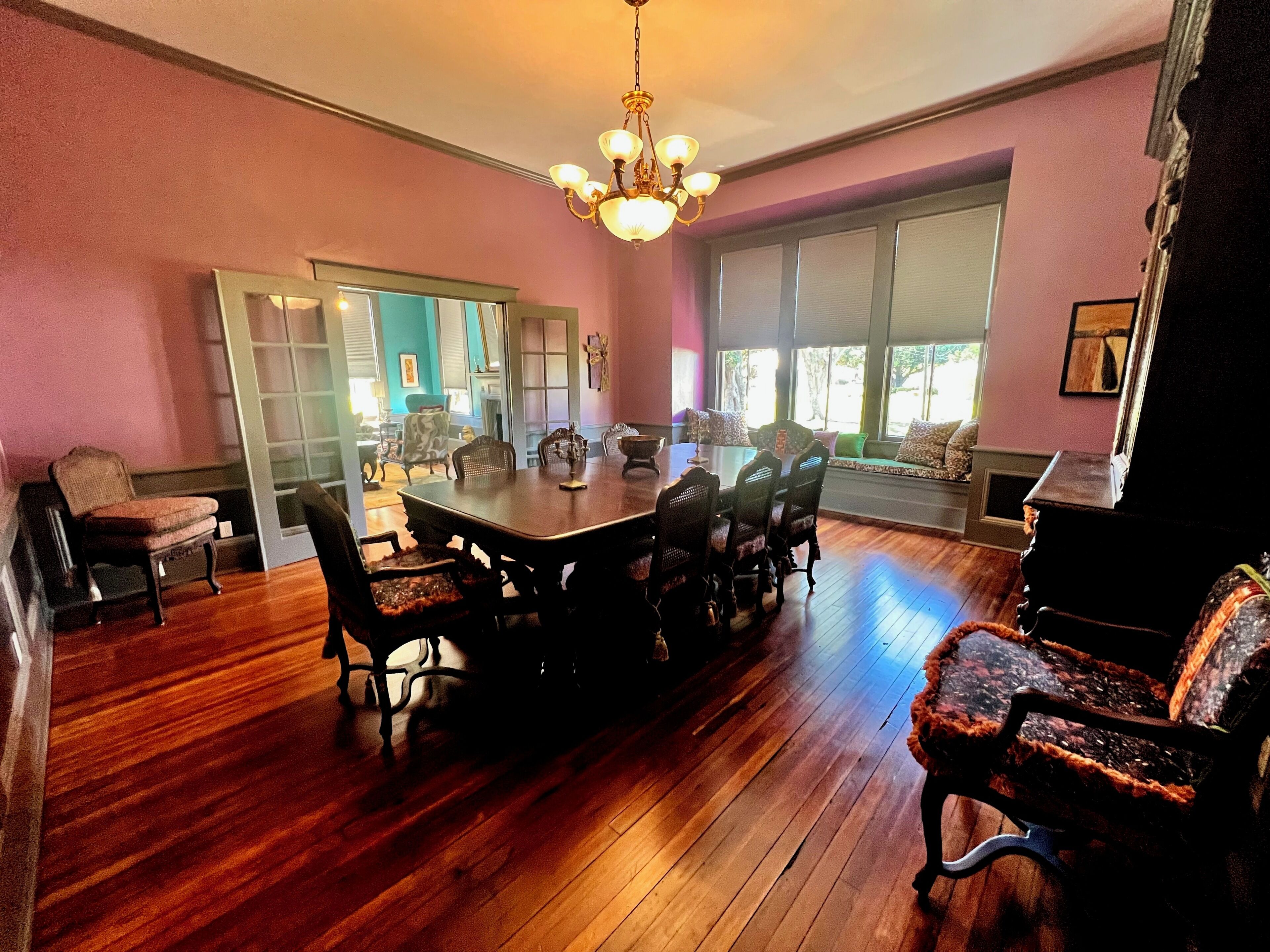 A dining area with a wooden table and chairs, a chandelier, and a view through a window.