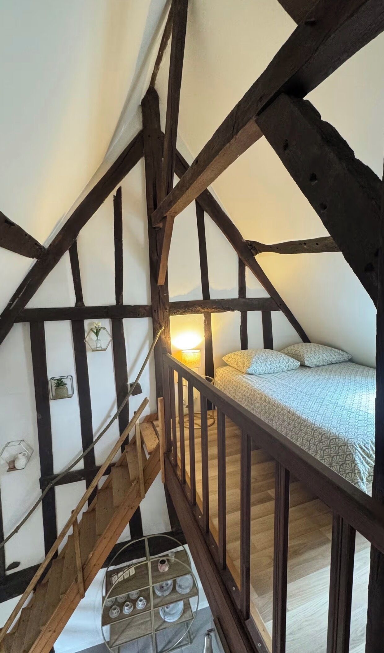A bedroom with a sloped ceiling, wooden beams, a bed with patterned bedding, a bedside lamp, and a small shelf with decorative items.