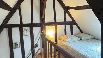 A bedroom with a sloped ceiling, wooden beams, a bed with patterned bedding, a bedside lamp, and a small shelf with decorative items.