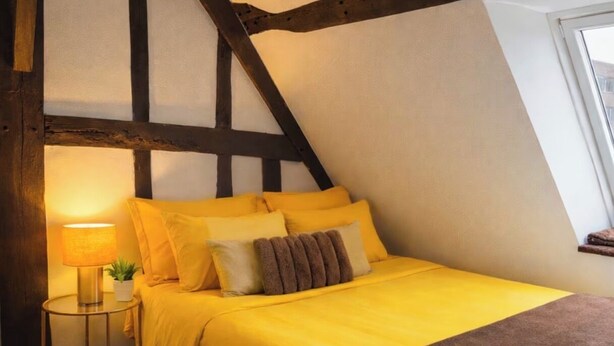 Un dormitorio con techo inclinado, una cama con ropa de cama amarilla, una lámpara de noche y una ventana.