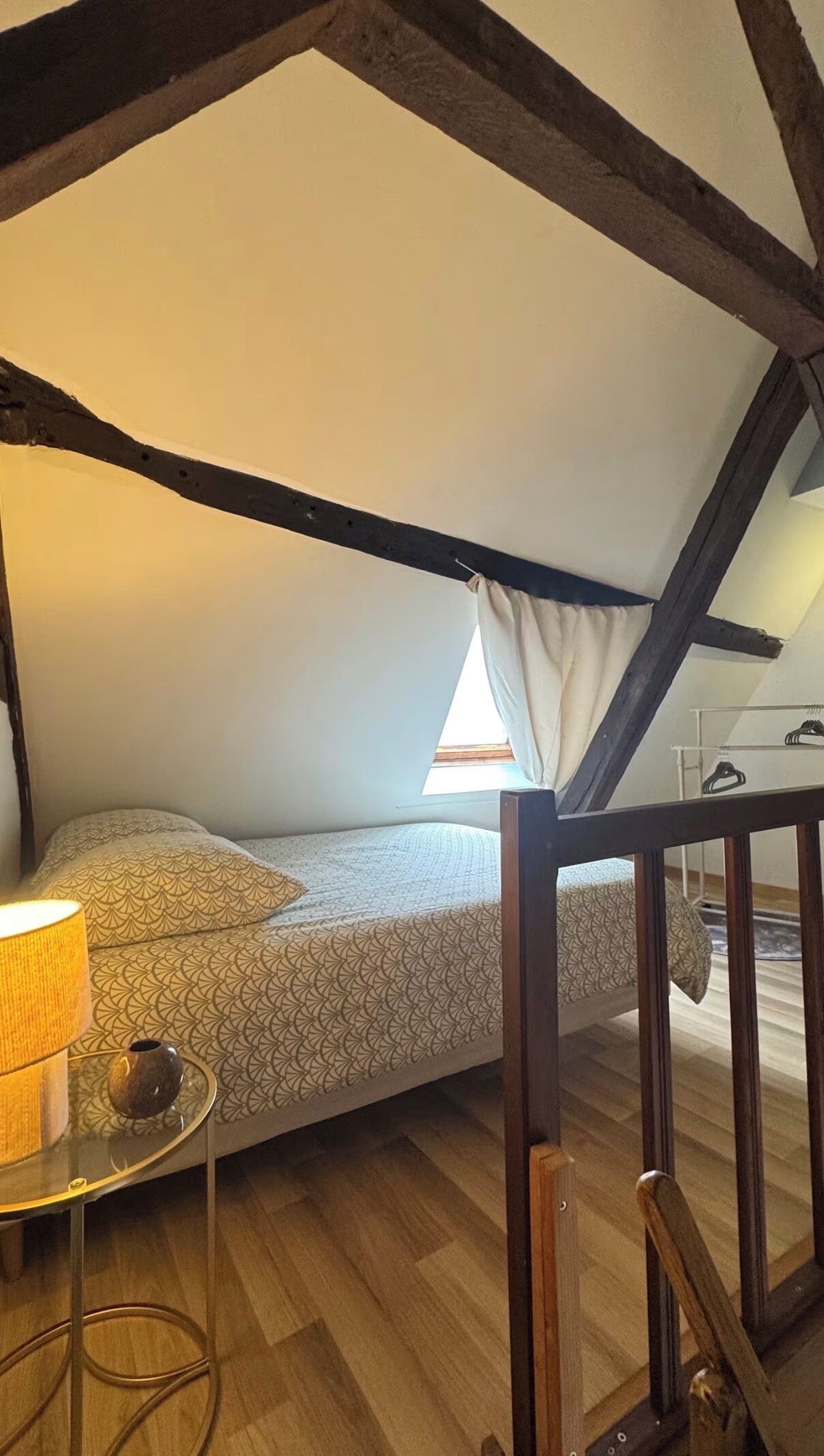 A small, cozy attic bedroom with a sloped ceiling, a single bed, a bedside table with a lamp, and a window with curtains.