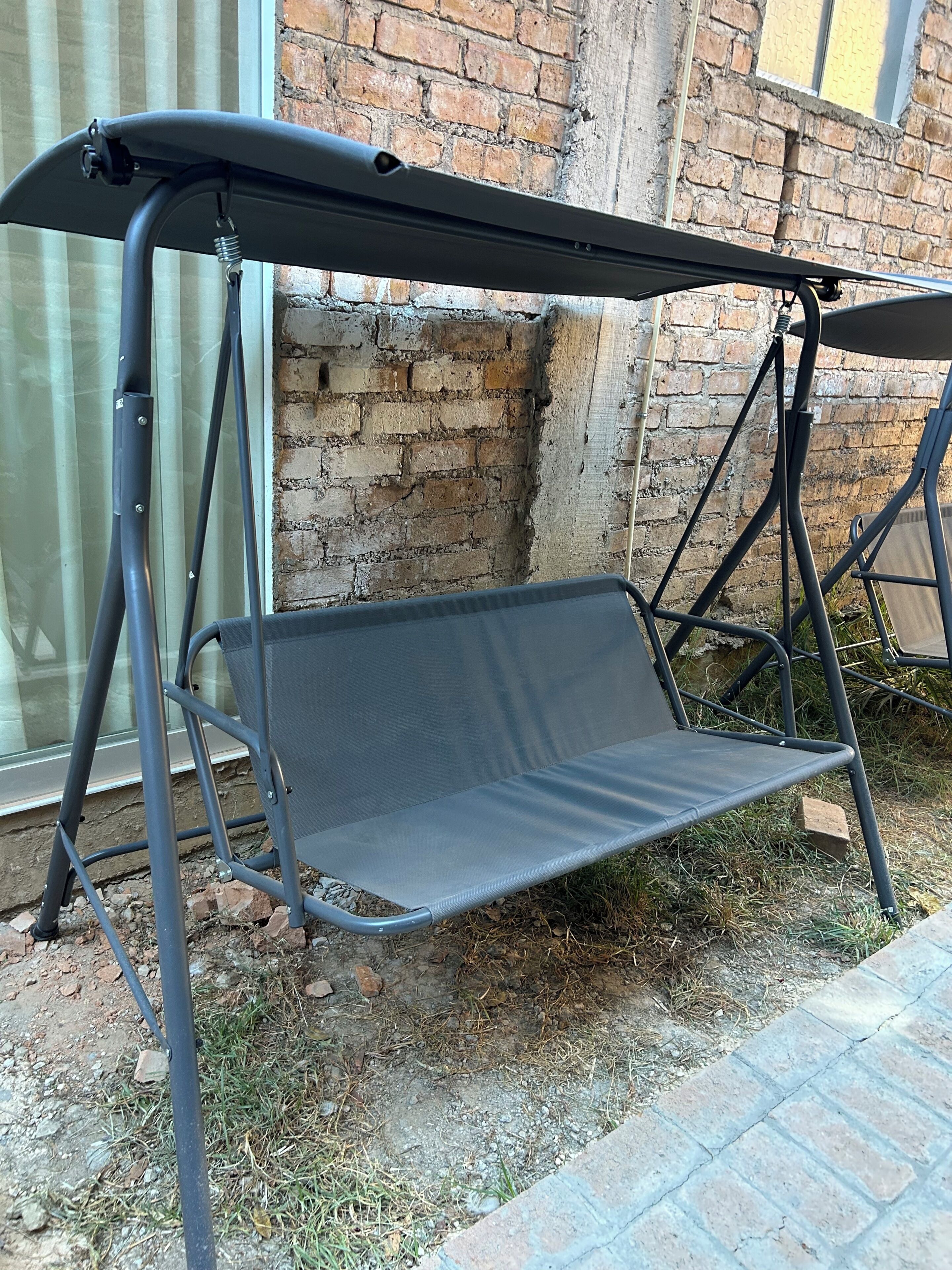 A metal swing chair with a canopy, positioned outdoors against a brick wall.