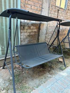 Una silla colgante de metal con dosel, ubicada al aire libre frente a un muro de ladrillo.