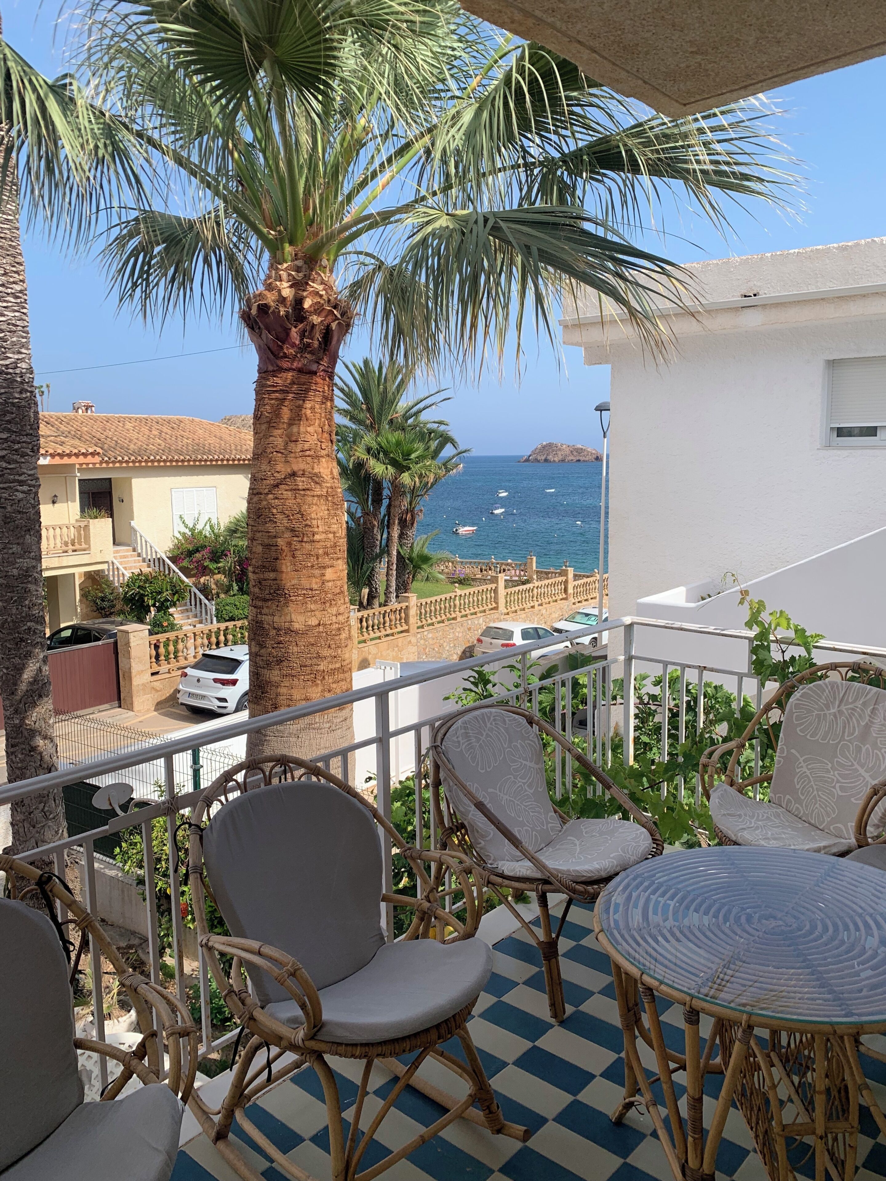 A balcony with wicker furniture, a palm tree, and a view of the sea and buildings.