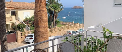A balcony with wicker furniture, a palm tree, and a view of the sea and buildings.