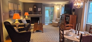 A living room with a sofa, two lamps, a fireplace, a television, and a dining area with a table set for two. - Greenbelt Getaway 3 Bedroom Home in Oak Ridge TN (Oak Ridge)