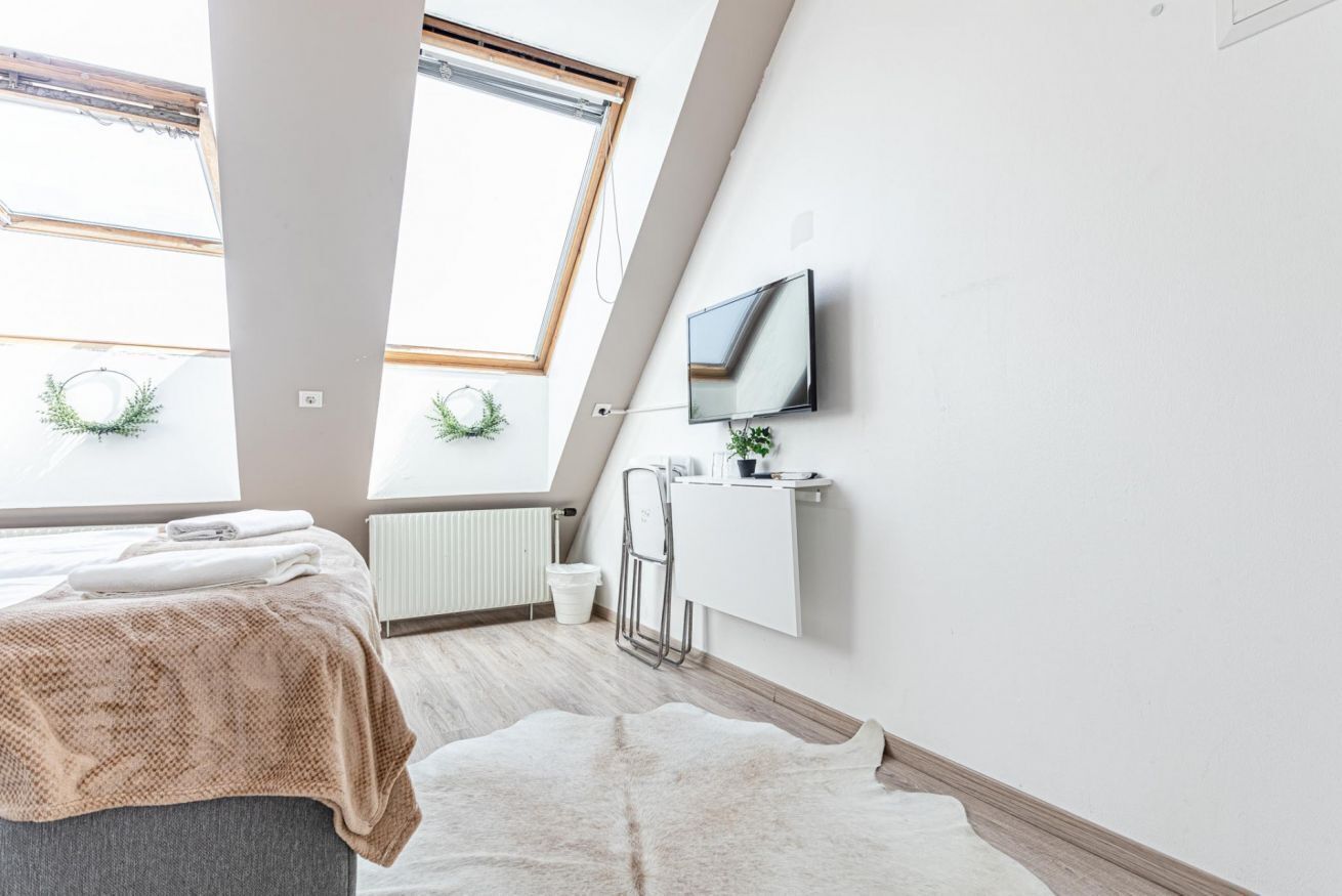 Une chambre moderne avec un plafond en pente, un lit recouvert d’une couverture blanche et marron, un téléviseur fixé au mur et un radiateur.