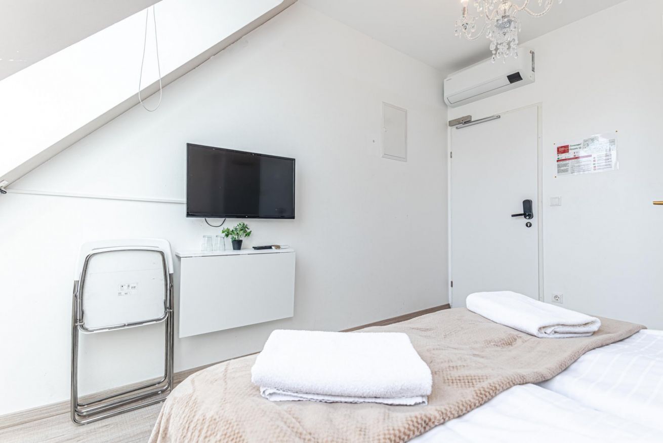 A modern hotel room with a flat-screen TV mounted on the wall, a small potted plant, a white bedside table, a folded white towel, and a door with a sign.