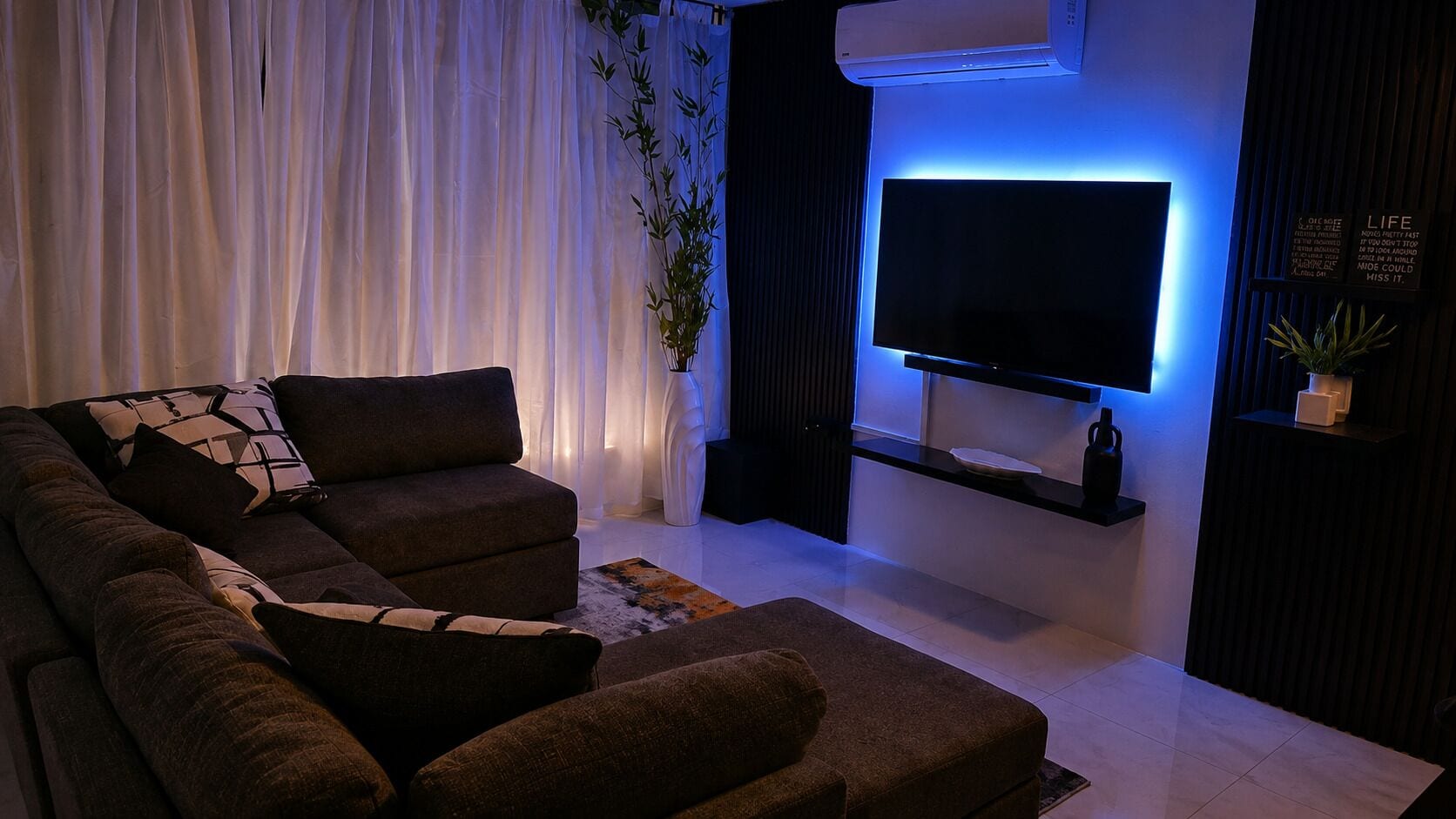 Un salon moderne avec un canapé d’angle, un téléviseur à écran plat fixé au mur et une plante décorative.