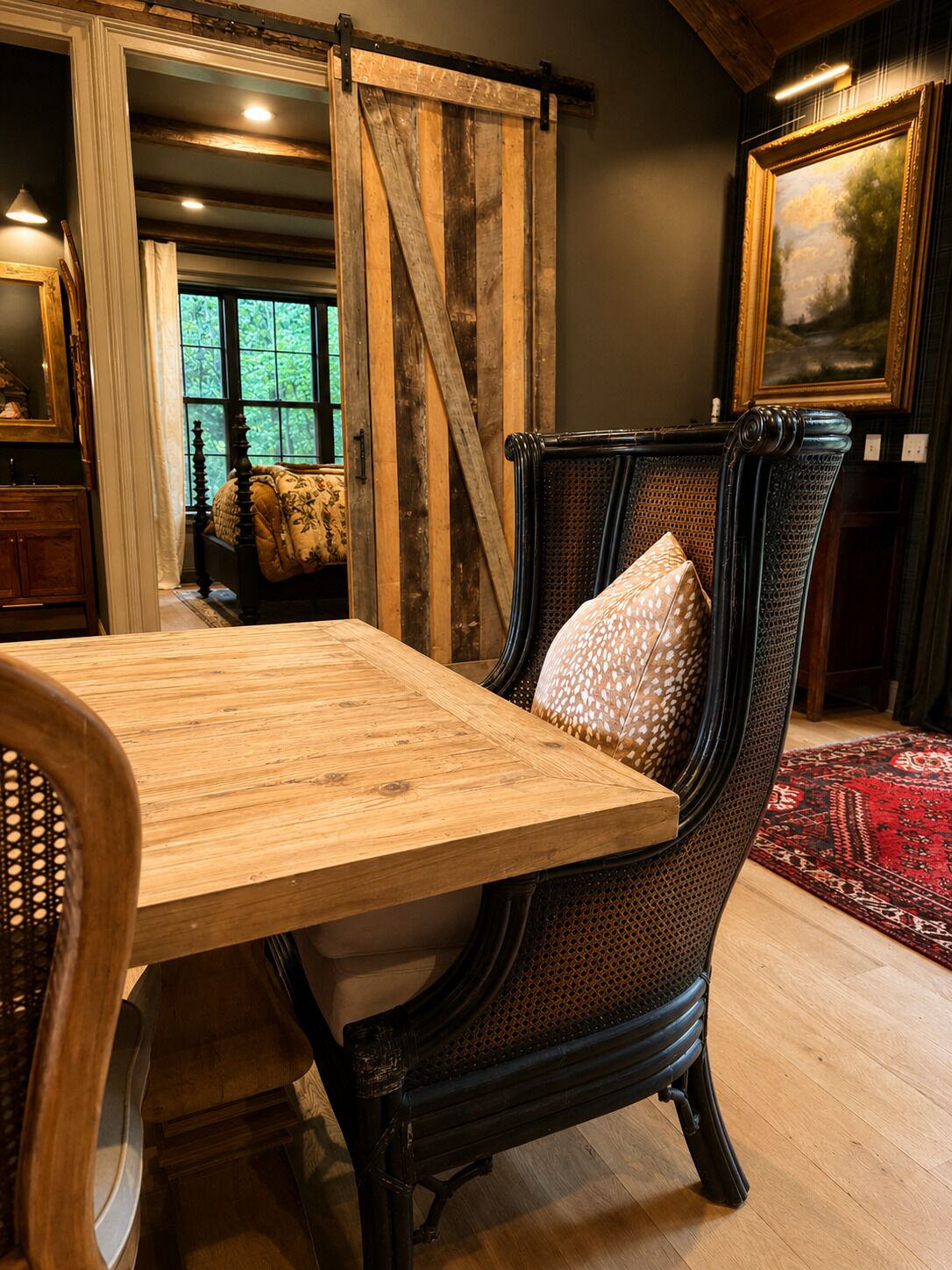 A rustic interior with a wooden dining table, a woven chair with a patterned cushion, and a large painting on the wall.