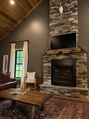 A living room with a stone fireplace, a mounted television, a wooden bench, and a patterned rug. - Luxury Private Mountain Spa - Trout Fishing + Trails (Copper Hill)