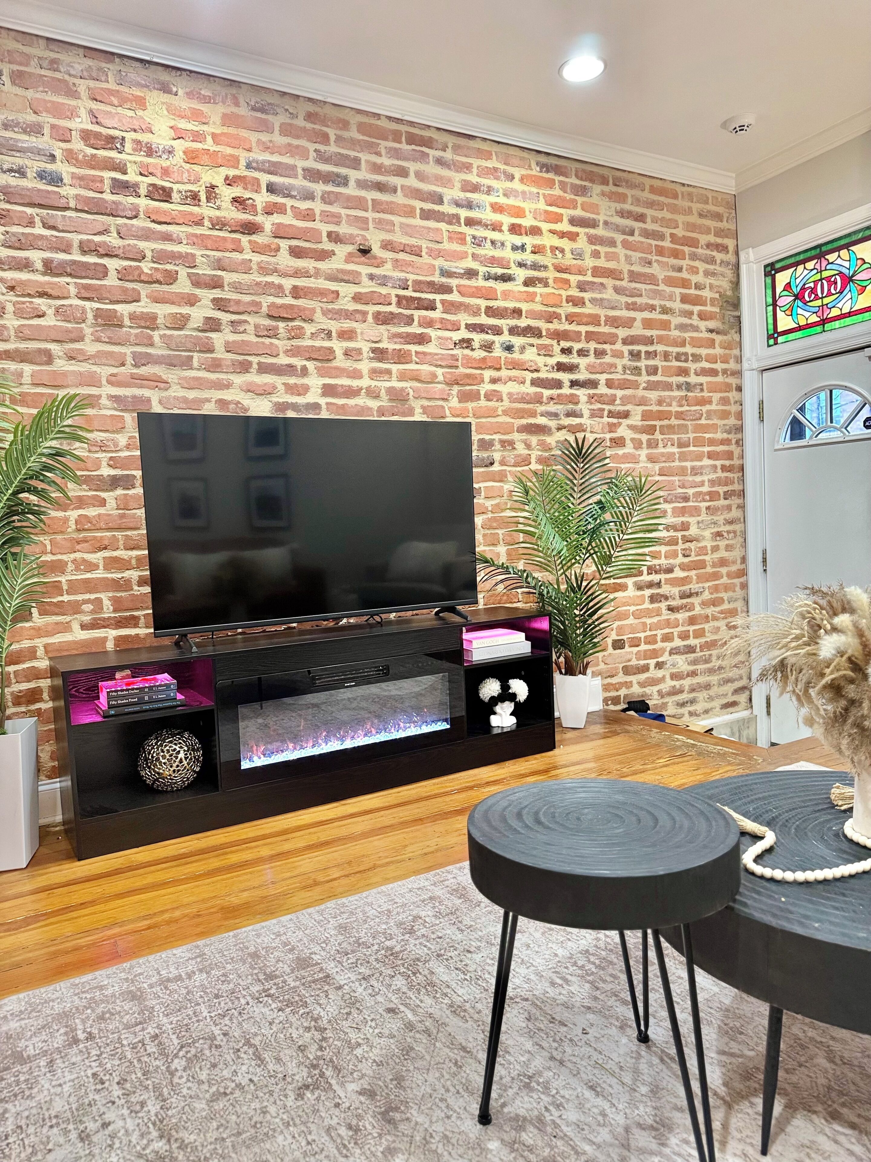Un salon avec un mur en briques, un téléviseur à écran plat posé sur un meuble noir, une table ronde noire et deux plantes en pot.