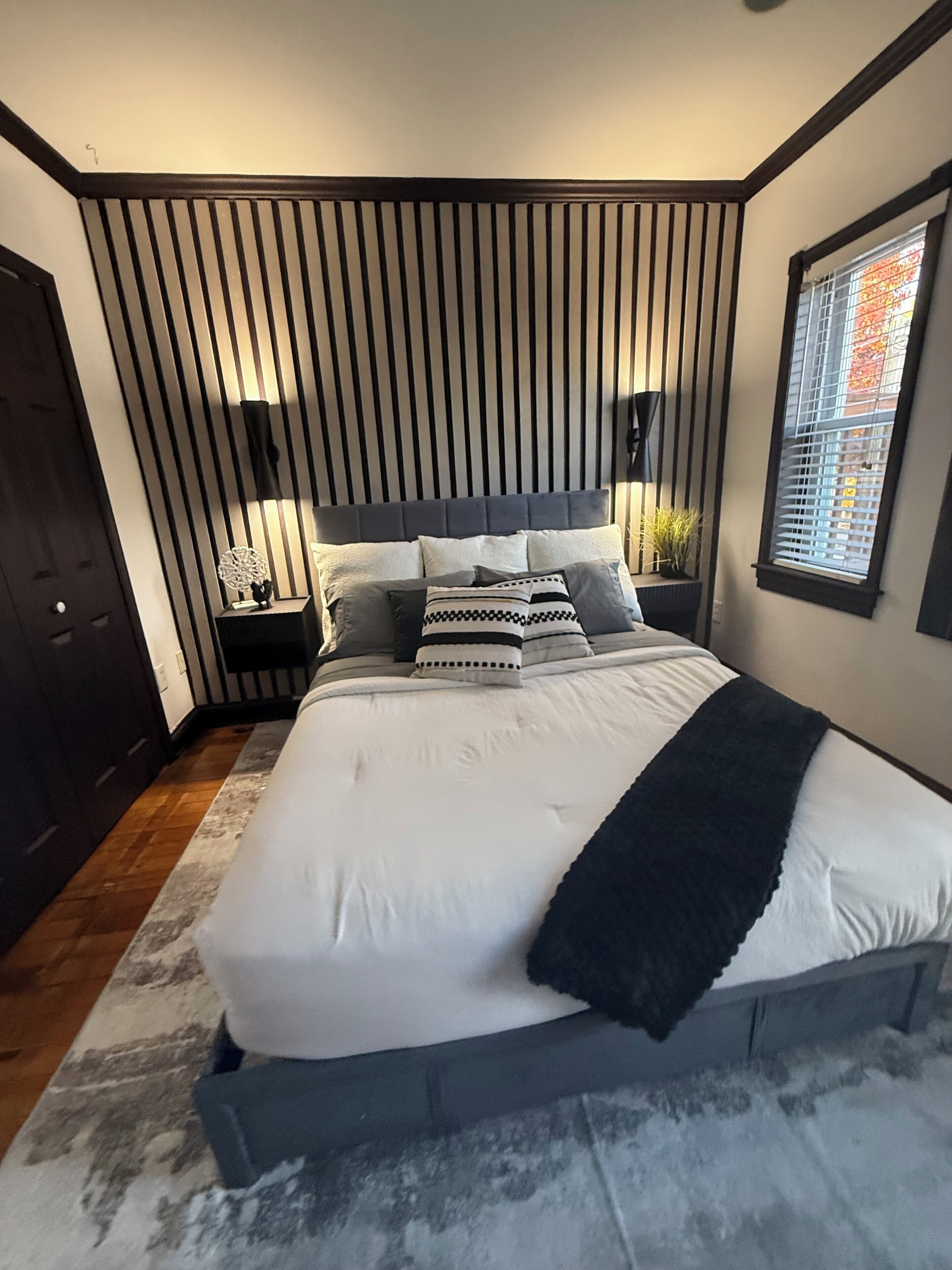 A modern bedroom with a large bed, a slatted wall design, and a window with blinds.