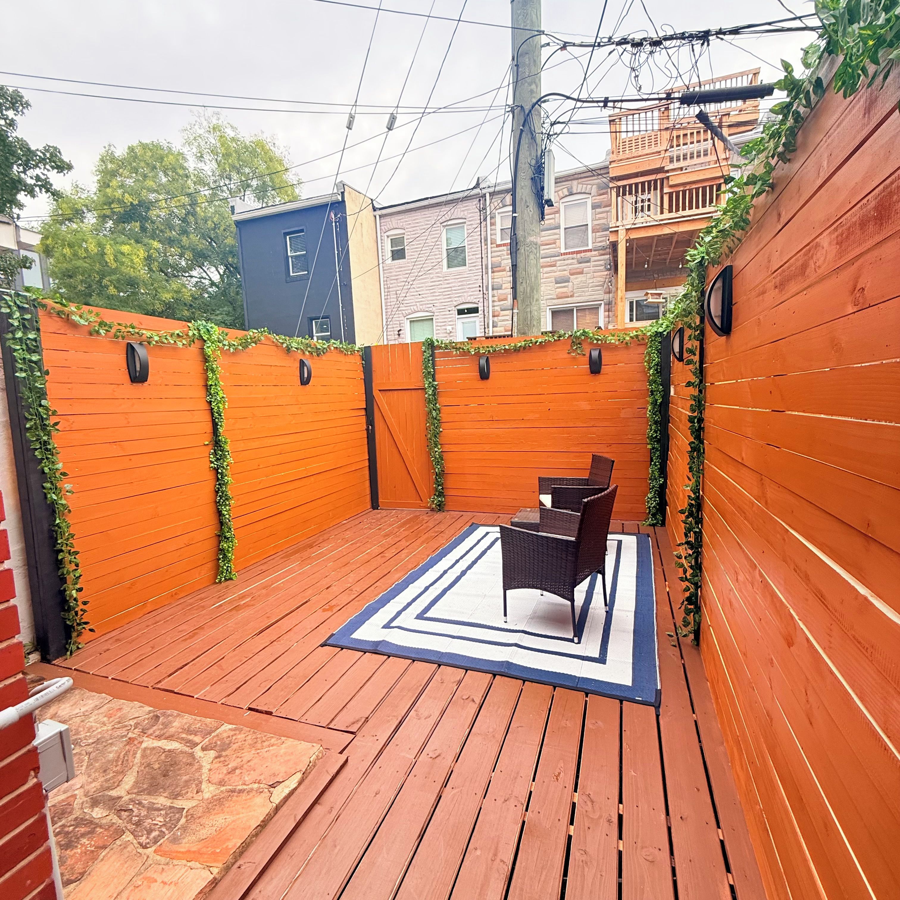 Una terraza de madera con un tapete azul y blanco, dos sillas de mimbre y una pared de ladrillo con plantas.