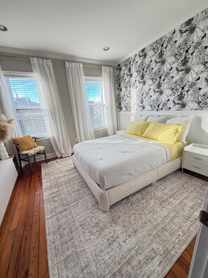 A bedroom with a large bed, a nightstand, a chair, and a window with curtains. - Cozy Canton Stay (Baltimore)