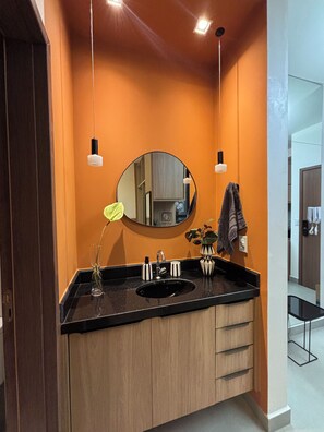 A bathroom with a black countertop, a round mirror, and wooden cabinets. - Monte Olympus: Apt moderno com sacada 10º andar (Uberlândia)