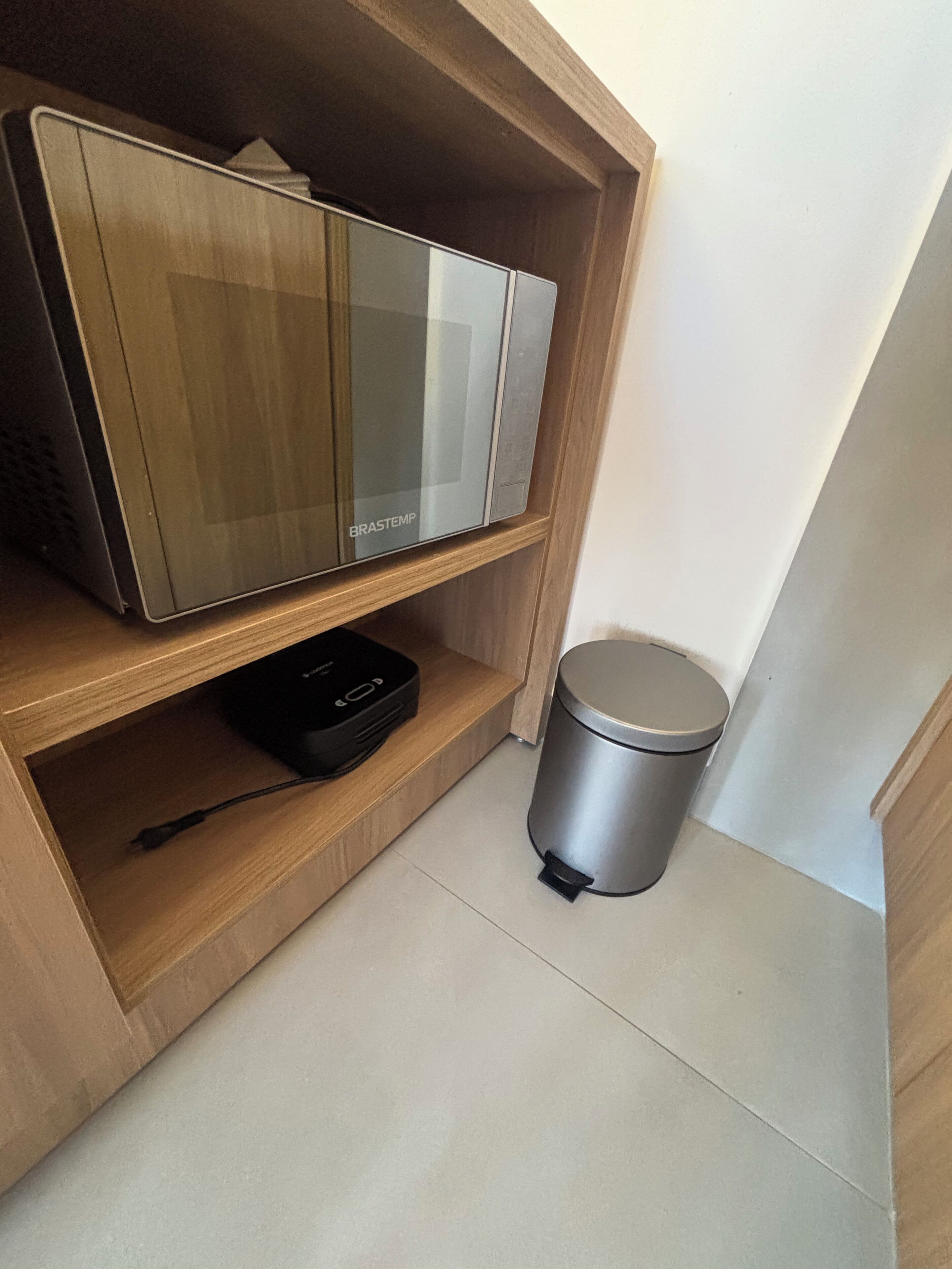 A kitchen area with a microwave, a trash bin, and a power strip on a wooden shelf.