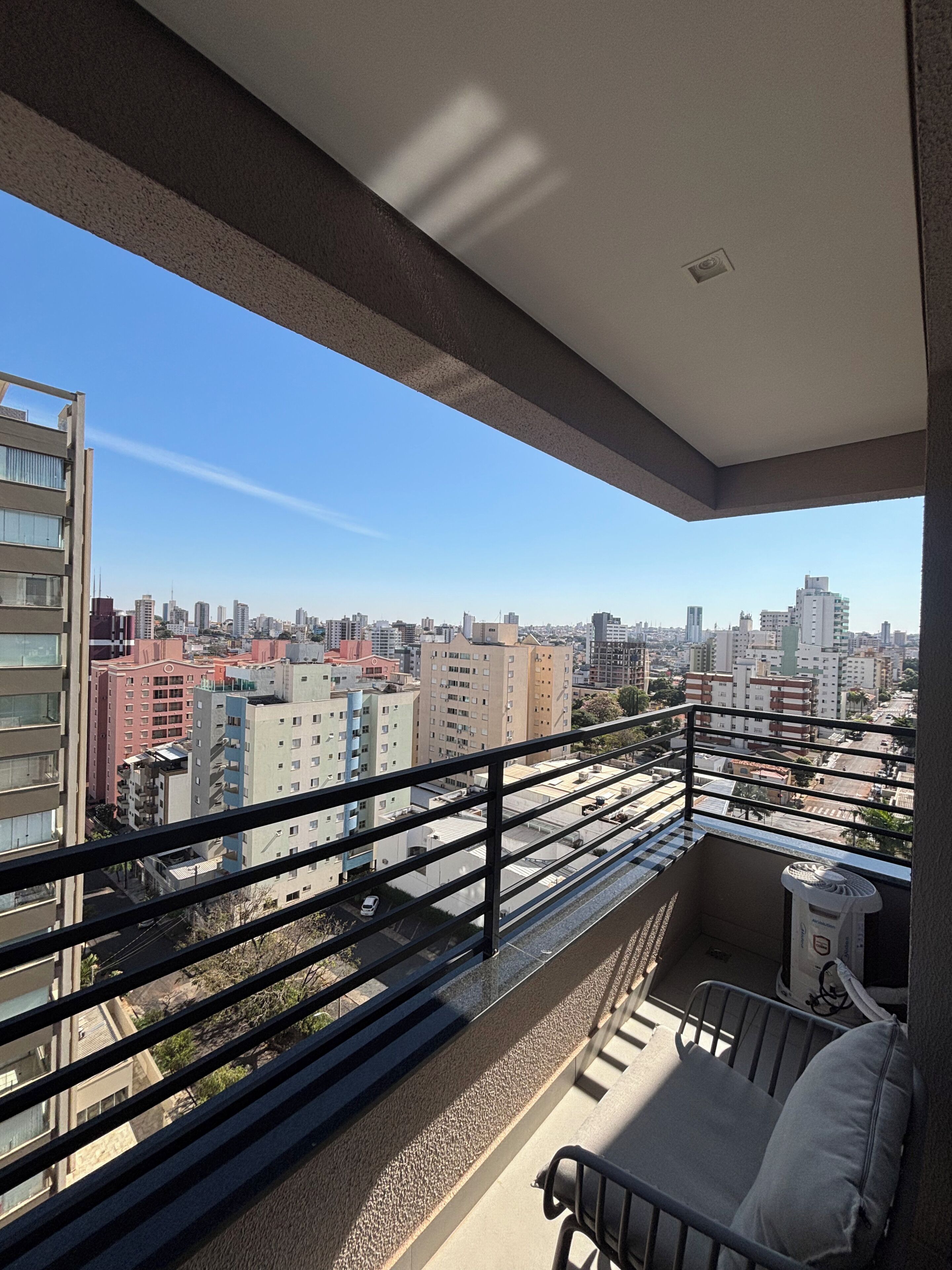 Un balcón con barandilla metálica, una pequeña zona de estar con cojines y vistas a un paisaje urbano con diversos edificios.