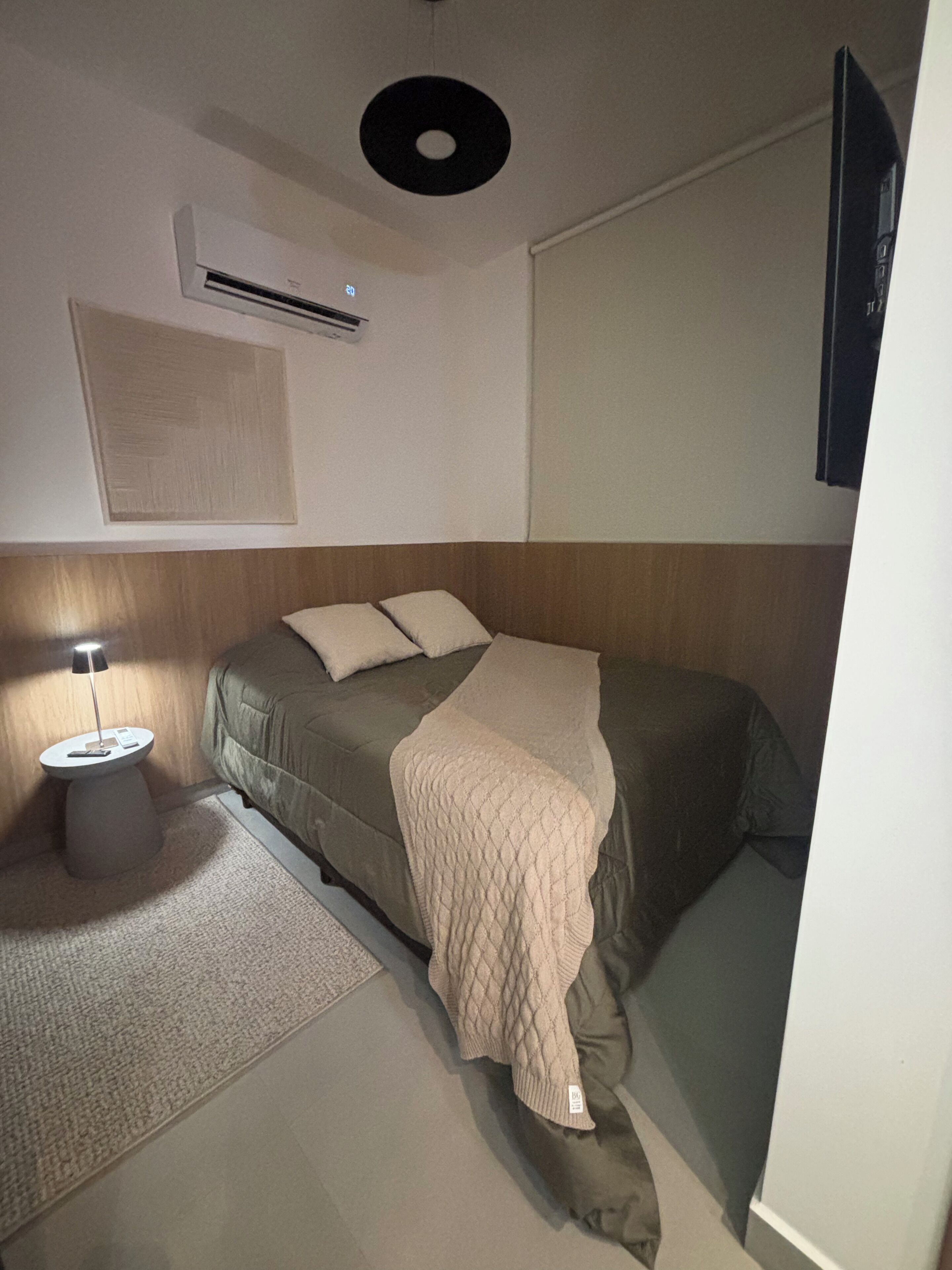 Habitación de hotel con cama, mesita de noche, lámpara, equipo de aire acondicionado y televisor montado en la pared.