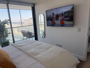 A hotel room with a large bed, a TV mounted on the wall, a view of the city through the window, and a small seating area with a chair and table. - Modern apt with king bed, sofa bed, washer/dryer, terrace. Near Parque Arauco. (Las Condes)