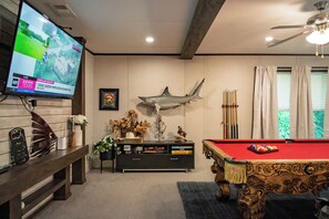 A living room with a pool table, a mounted television displaying a news channel, a decorative shark on the wall, and a billiards rack. - 4 bed, 2 bath home on the Brazos River and less than 4 miles from Lake Whitney. (Clifton)