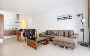 Ein modernes Wohnzimmer mit einer Couch, einem Couchtisch und Sesseln. Eine Küchenzeile mit Schränken und einer Mikrowelle ist ebenfalls vorhanden. An der Wand hängt ein Gemälde, und ein Stehschrank ist sichtbar.