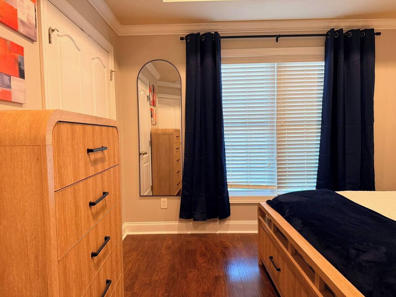 Un dormitorio con un aparador de madera, una cama con una manta oscura, una ventana con persianas y un espejo en la pared.