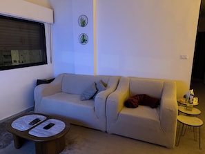 A living room with a sofa, two side tables, a window, and wall art. - 3-bedroom apartment prime location (Amman)