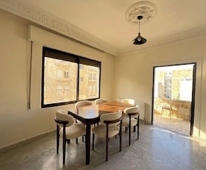 A dining room with a wooden table and six chairs, large windows with views of buildings, and a ceiling light fixture. - 3-bedroom apartment prime location (Amman)