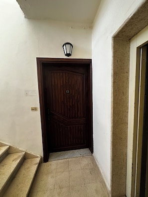 A wooden door with a light fixture above it, a set of stairs leading up to the door, and a wall with a textured finish. - 3-bedroom apartment prime location (Amman)
