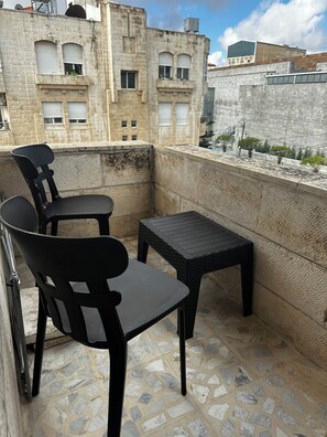 A balcony with two chairs and a small table, stone flooring, and a view of a building with windows and a wall. - 3-bedroom apartment prime location (Amman)