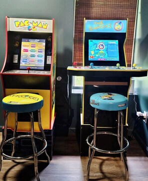 Dos máquinas de videojuegos, una con la etiqueta 'PAC-MAN' y la otra 'THE SIMPSONS', con taburetes delante de cada una.