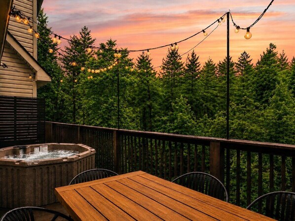 An outdoor deck with a hot tub, wooden table, and chairs, surrounded by trees and string lights against a sunset sky.