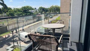 A balcony with two wicker chairs and a small table, overlooking a street with vehicles and greenery. - Studio Apartment  (Accra)