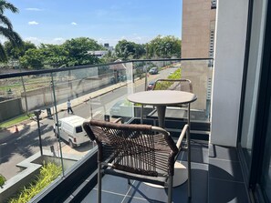 A balcony with two wicker chairs and a small table, overlooking a street with vehicles and greenery. - Studio Apartment  (Accra)