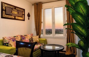 A living room with a green sofa, a round table, a large window with a view, and a decorative wall piece.