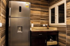 A stainless steel refrigerator with a water dispenser, a wooden backsplash, a window with a white frame, and a dark wooden cabinet with a tea kettle.